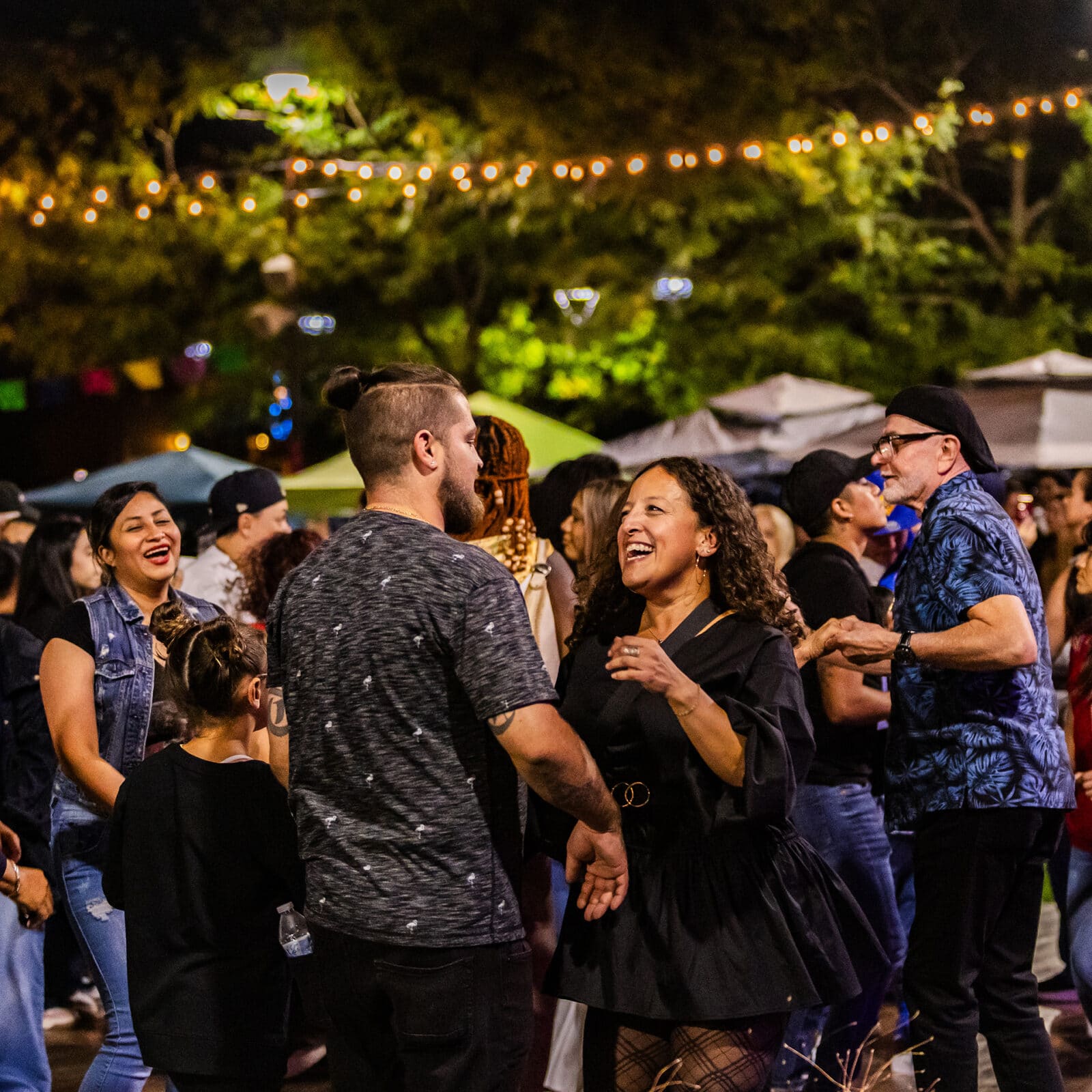 Photograph of a large crowd of people dancing during the 2024 Akron Latin Festival in downtown Akron. The photo is outdoors on a late summer night. Several people, of all ages, are smiling and laughing. Photograph by Talia Hodge