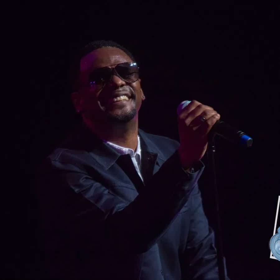 Photograph of American R&B singer Carl Thomas on a darkened stage, holding a microphone. Thomas is wearing stylish sunglasses and a blue sports jacket, and is smiling as he performs a song.