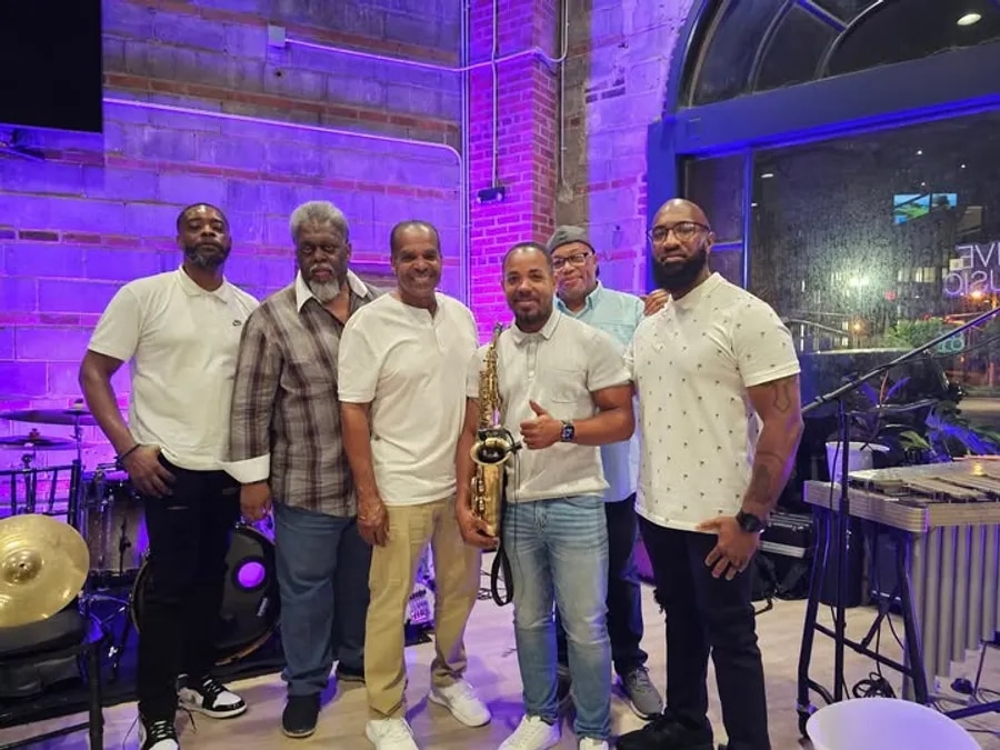 Photograph of 6 members of the contemporary jazz/R&B group FORECAST in a bar and grill setting. All 6 members are smiling at the camera, one member is holding a saxophone.